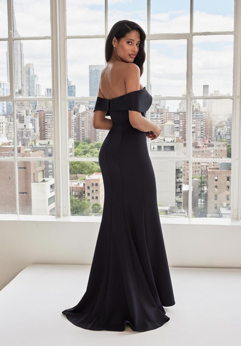 Woman in a black off-the-shoulder gown, smiling, near a window with city view.