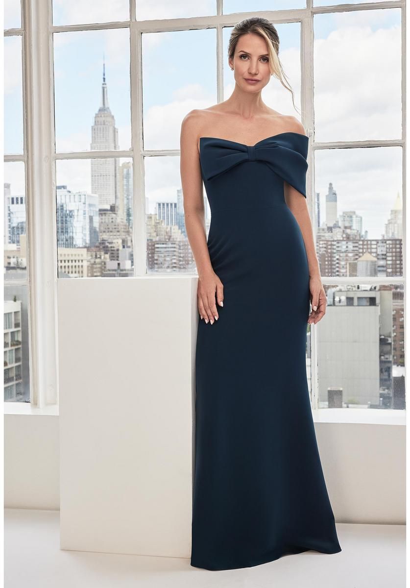 Woman in a dark blue off-the-shoulder gown, standing by a window with a city view.