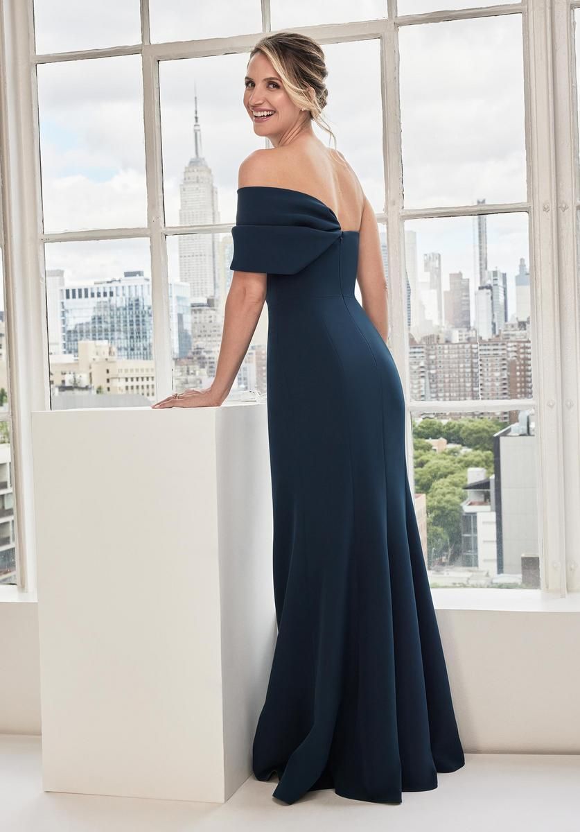 Woman in a dark blue off-the-shoulder gown, standing by a window with a city view.