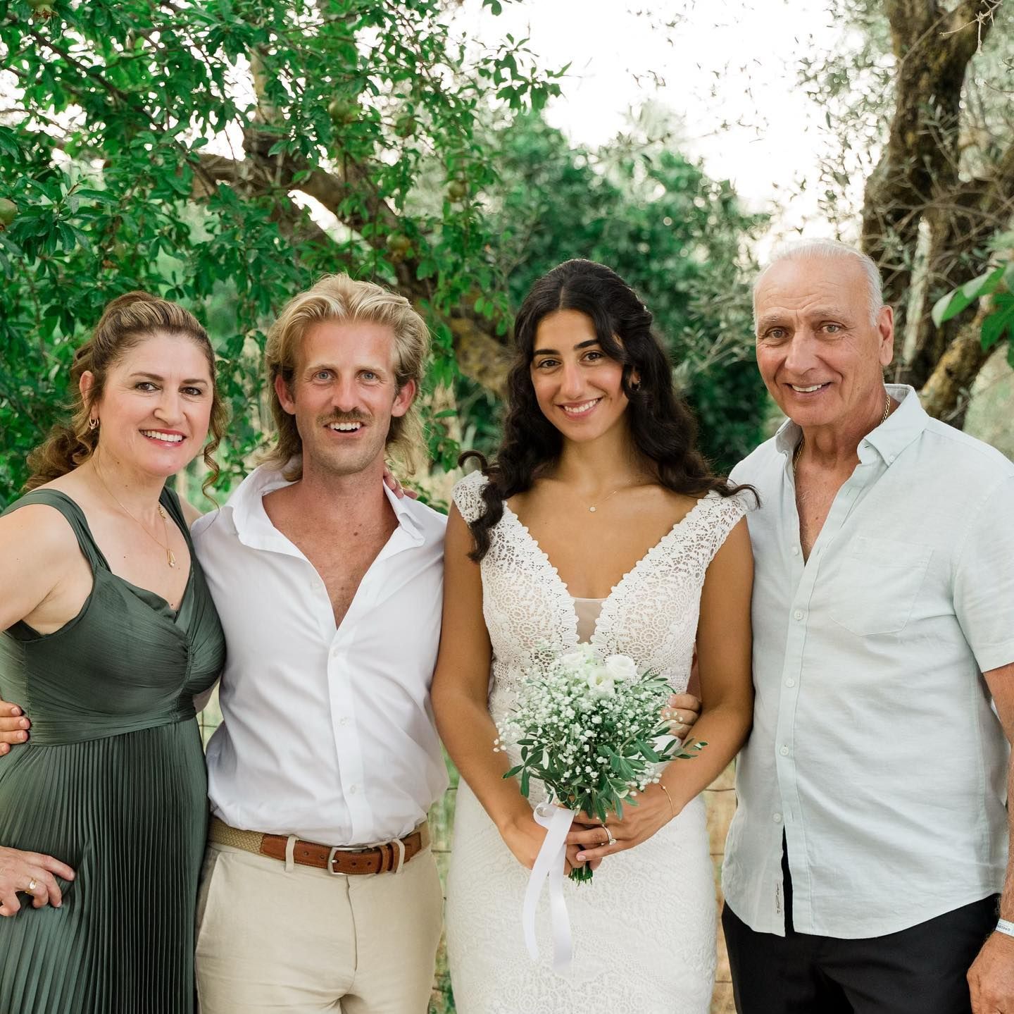 Wedding photo: Bride and groom with two others. Bride in white dress, holding bouquet. Others are smiling, outdoors.