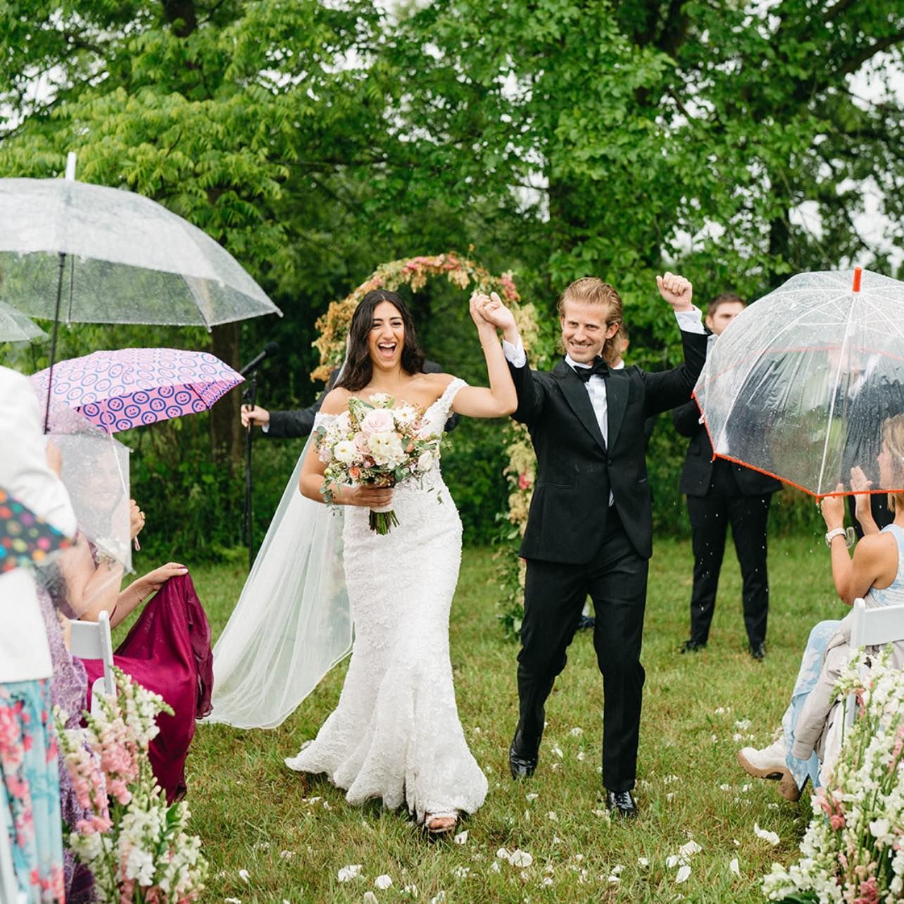 Bride and groom celebrate after a wedding ceremony under umbrellas on a grassy lawn.