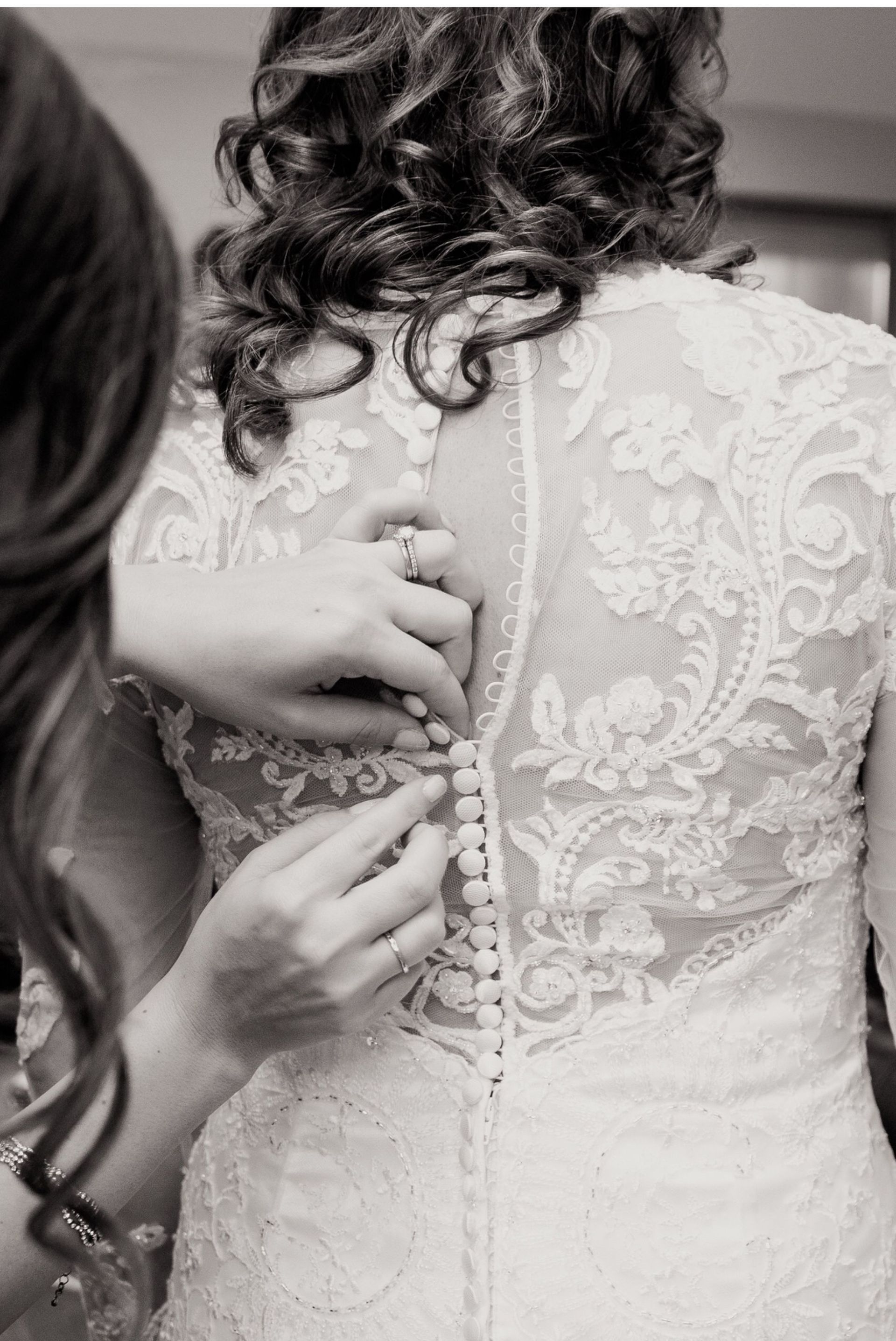 Woman's back in lace wedding dress; someone fastens buttons.