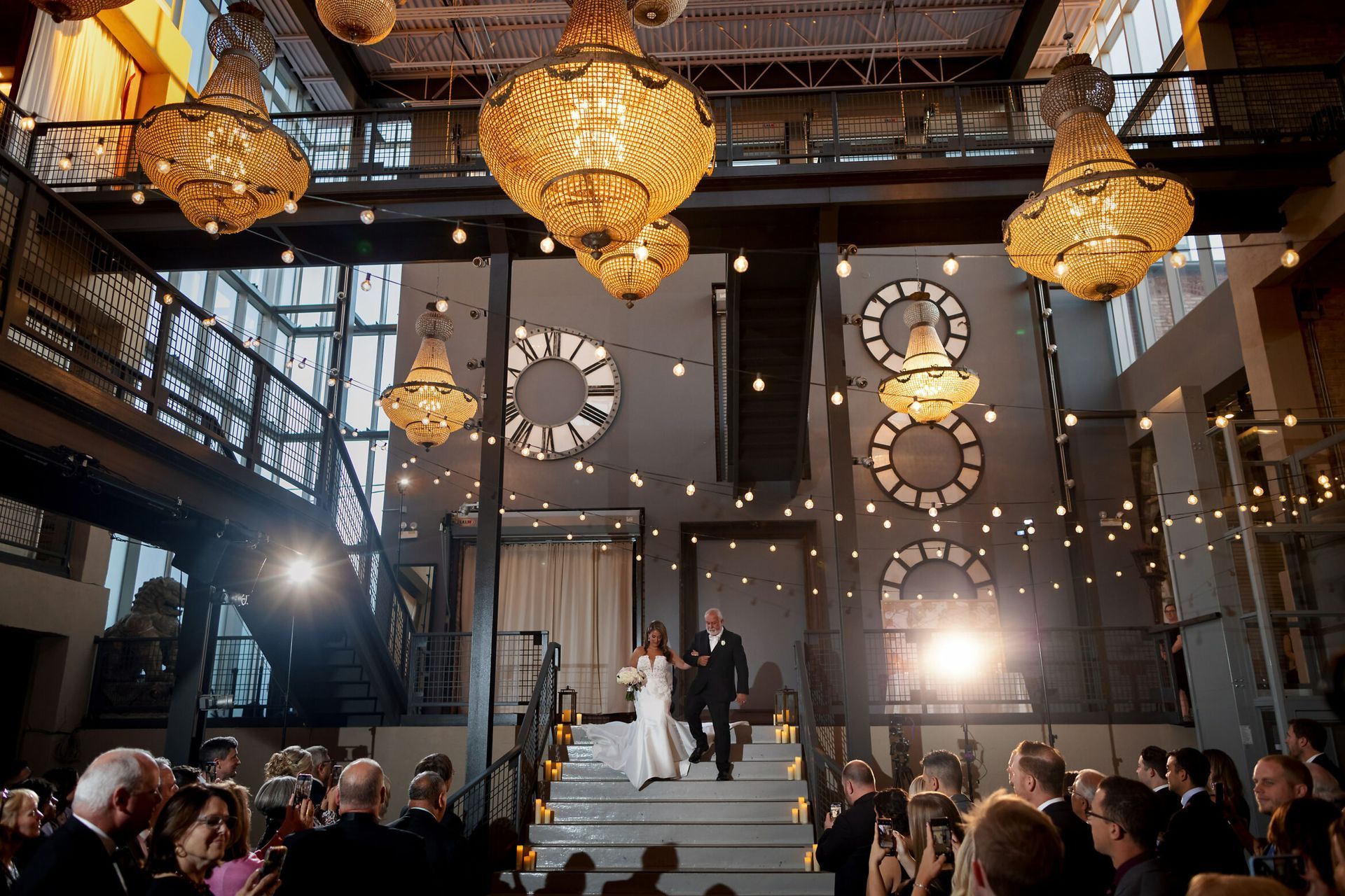 Wedding ceremony, bride and officiant descend stairs. Industrial setting with chandeliers, guests watch.