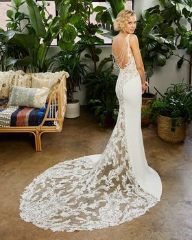Woman in a white wedding dress with lace train, standing in a room with plants.