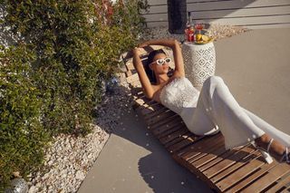 Woman in white jumpsuit and sunglasses relaxes on a wooden lounge chair by a wall, drinks nearby.