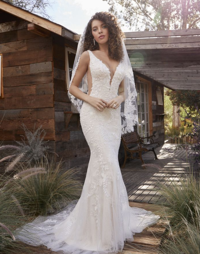 Woman in a white lace wedding dress with a deep V-neck, standing outside a wooden cabin with a veil.