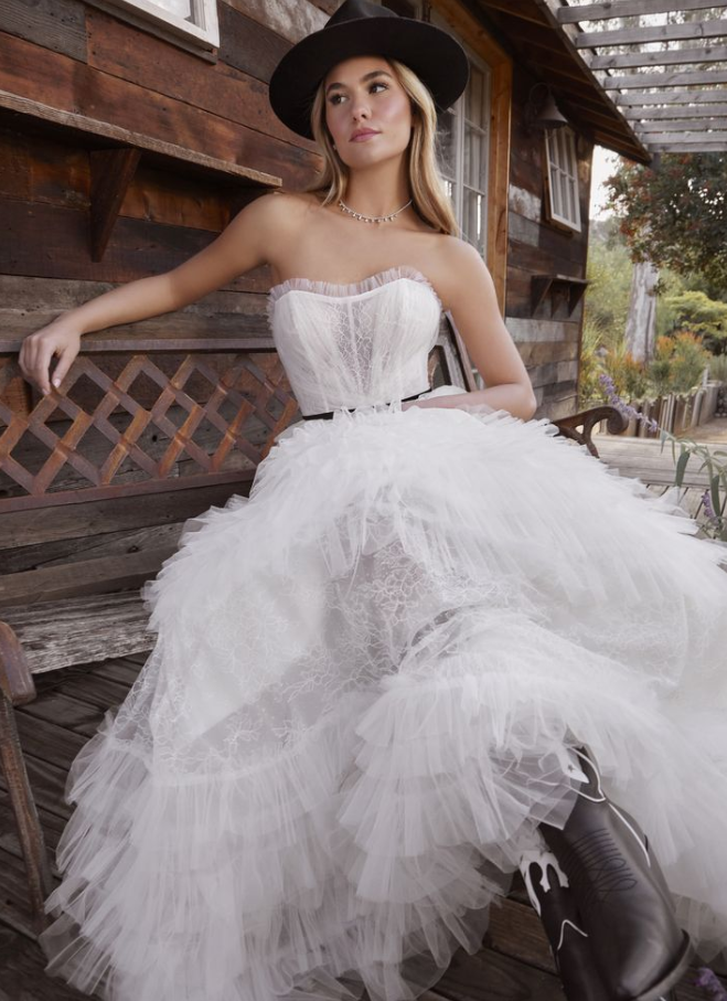 Woman in white strapless dress and cowboy boots, wearing a hat, sitting on a wooden bench.