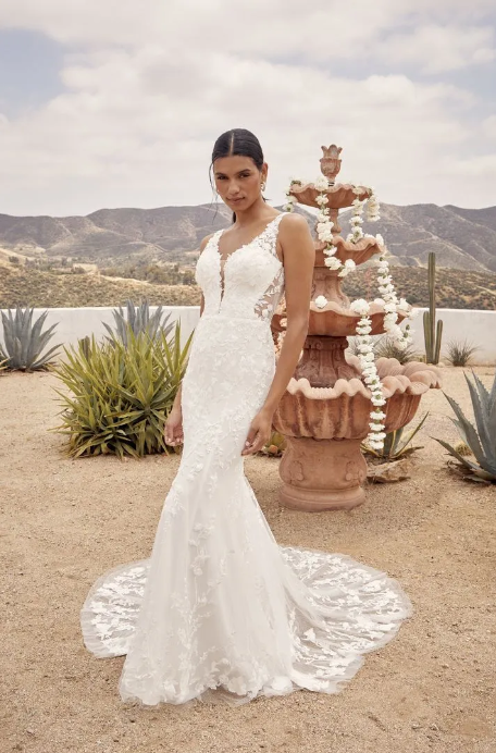 Woman in white wedding dress poses outdoors near a fountain, with a desert background.