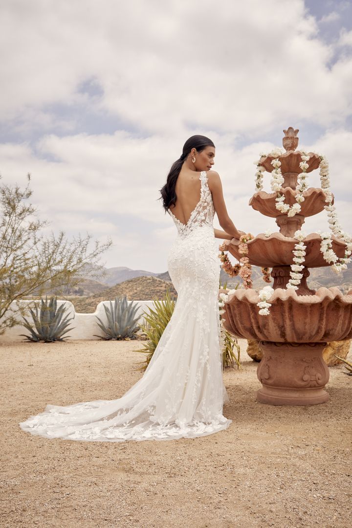 Woman in a white wedding dress stands near a fountain in an outdoor desert setting.