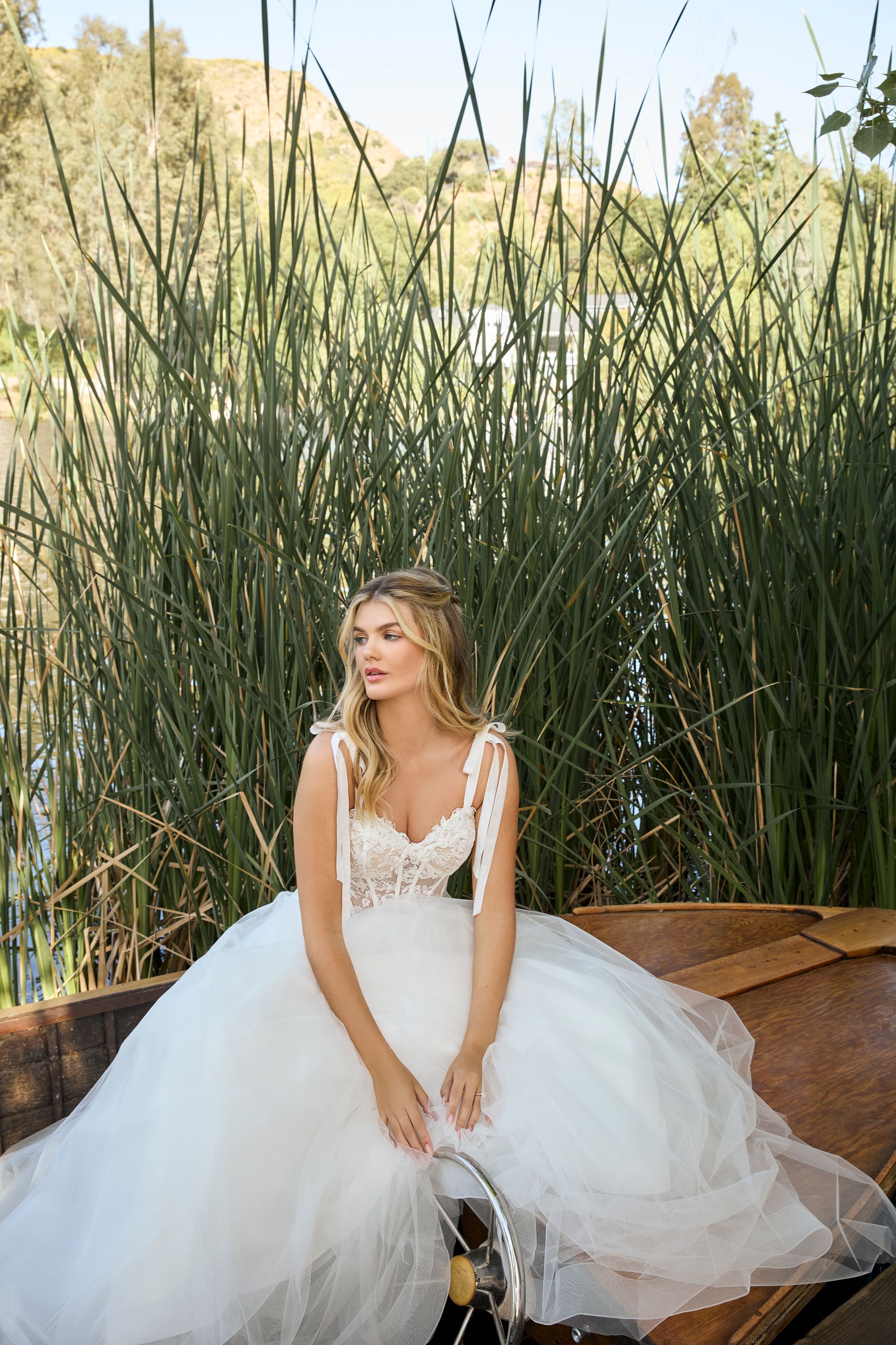Woman in a white wedding dress sits in a wooden boat, against a backdrop of tall grass and water.