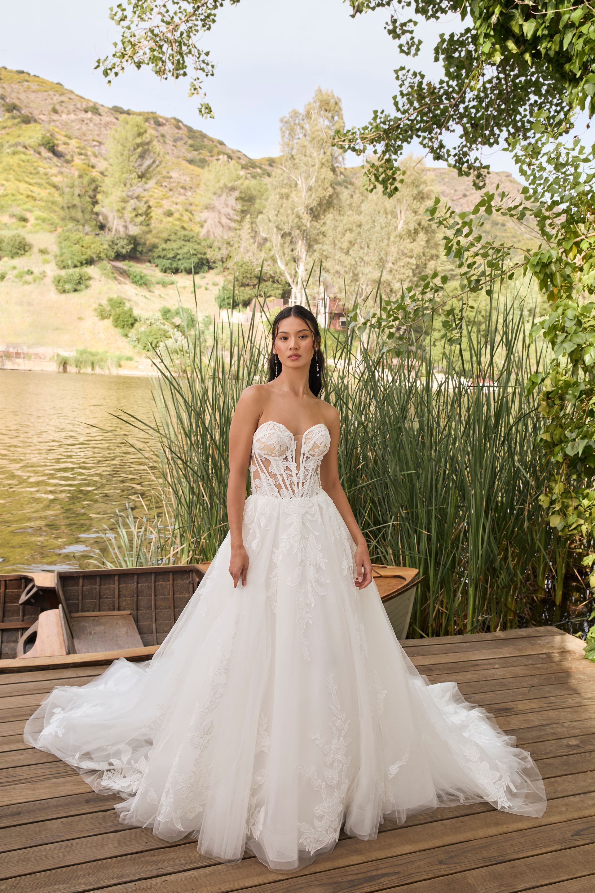 Woman in strapless white wedding dress stands by a lake, a boat in the background.