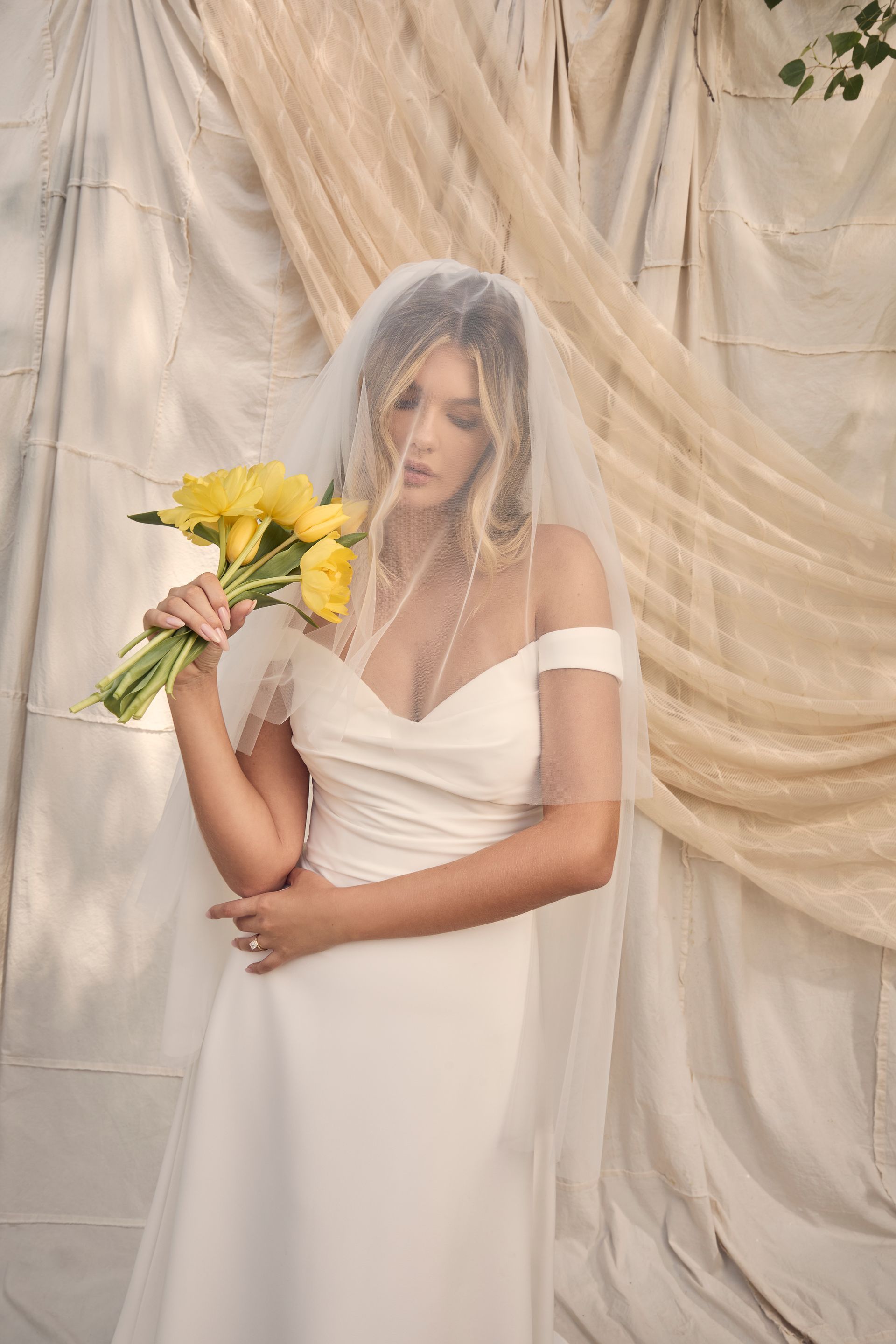Woman in wedding dress, veil, holding yellow flowers, draped backdrop.