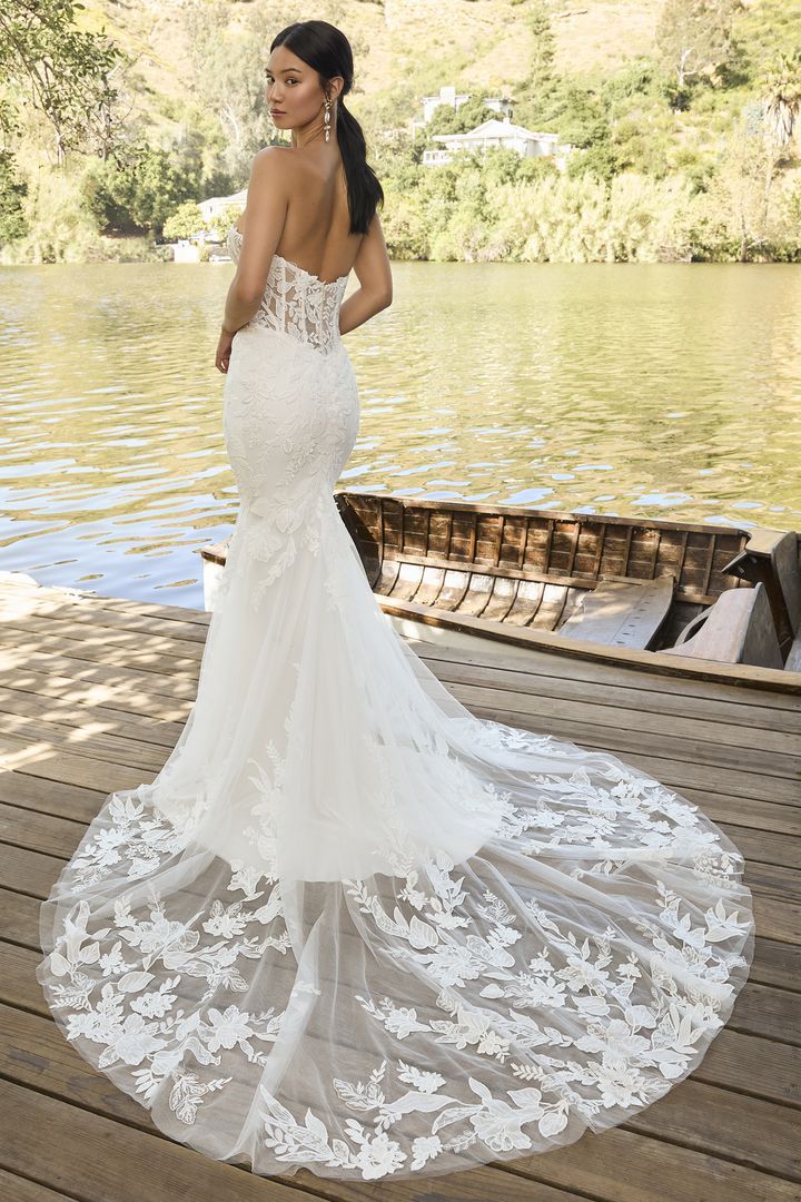 Woman in a white lace wedding gown with a long train, standing near water.