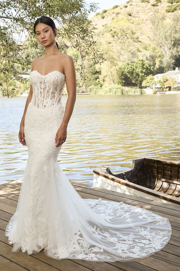 Woman in a white lace wedding gown with a long train, standing near water.