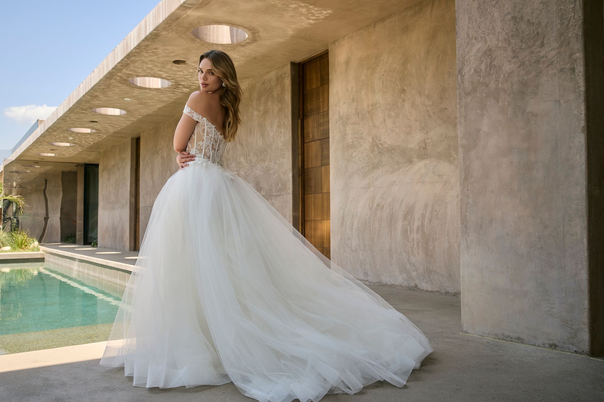 Woman in a white wedding gown, looking back, standing near a pool, in front of a modern building.
