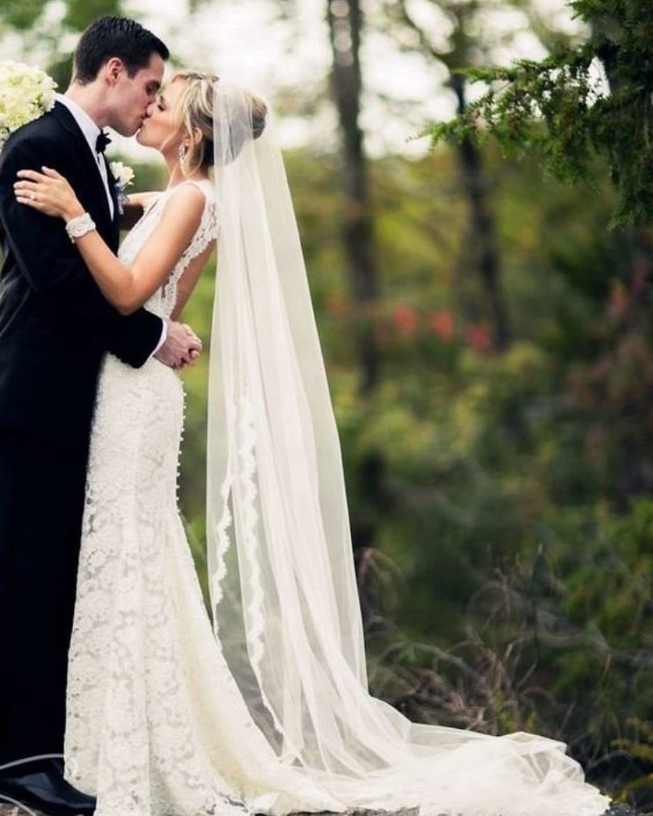 Bride and groom kissing outdoors; she wears a lace gown and veil, he wears a tuxedo.