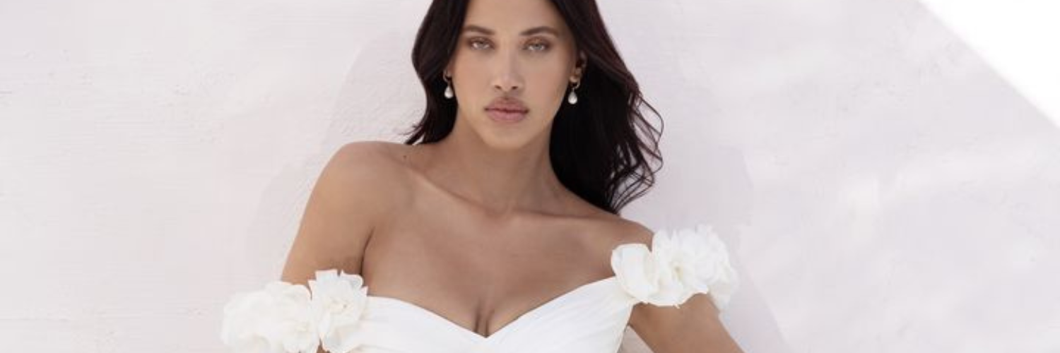 Woman in a white off-the-shoulder dress with white flowers. She looks at the viewer with dark hair.