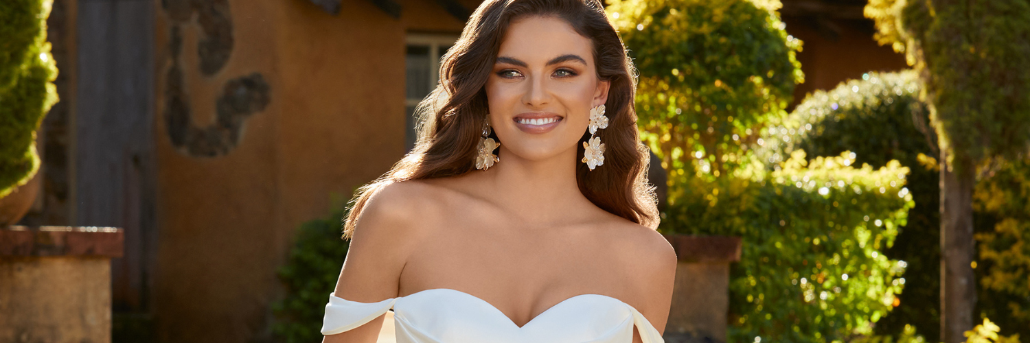 Woman in a white off-the-shoulder dress smiles outdoors, trees and building background.
