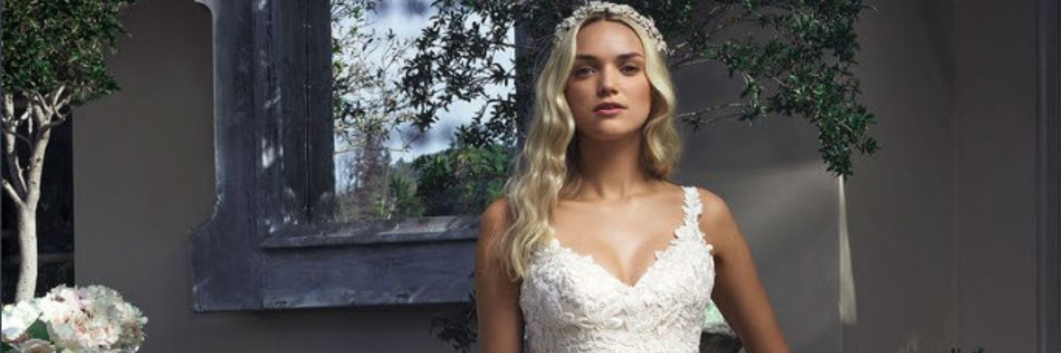 Blonde woman in a white wedding dress, standing outdoors. She is wearing a headpiece, and there is foliage in the background.