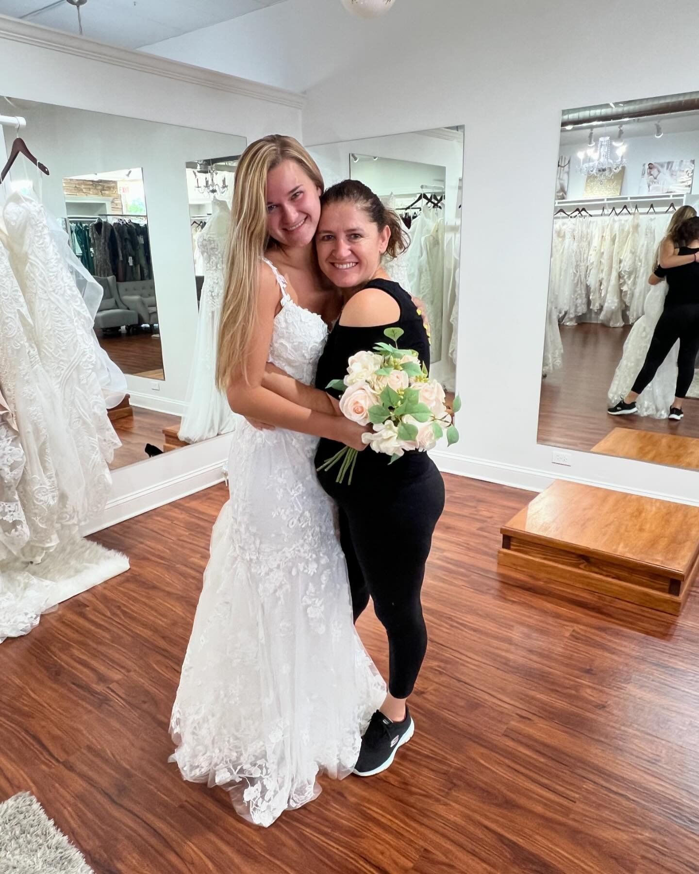 Woman in a white wedding dress hugs another woman holding a bouquet in a bridal shop.