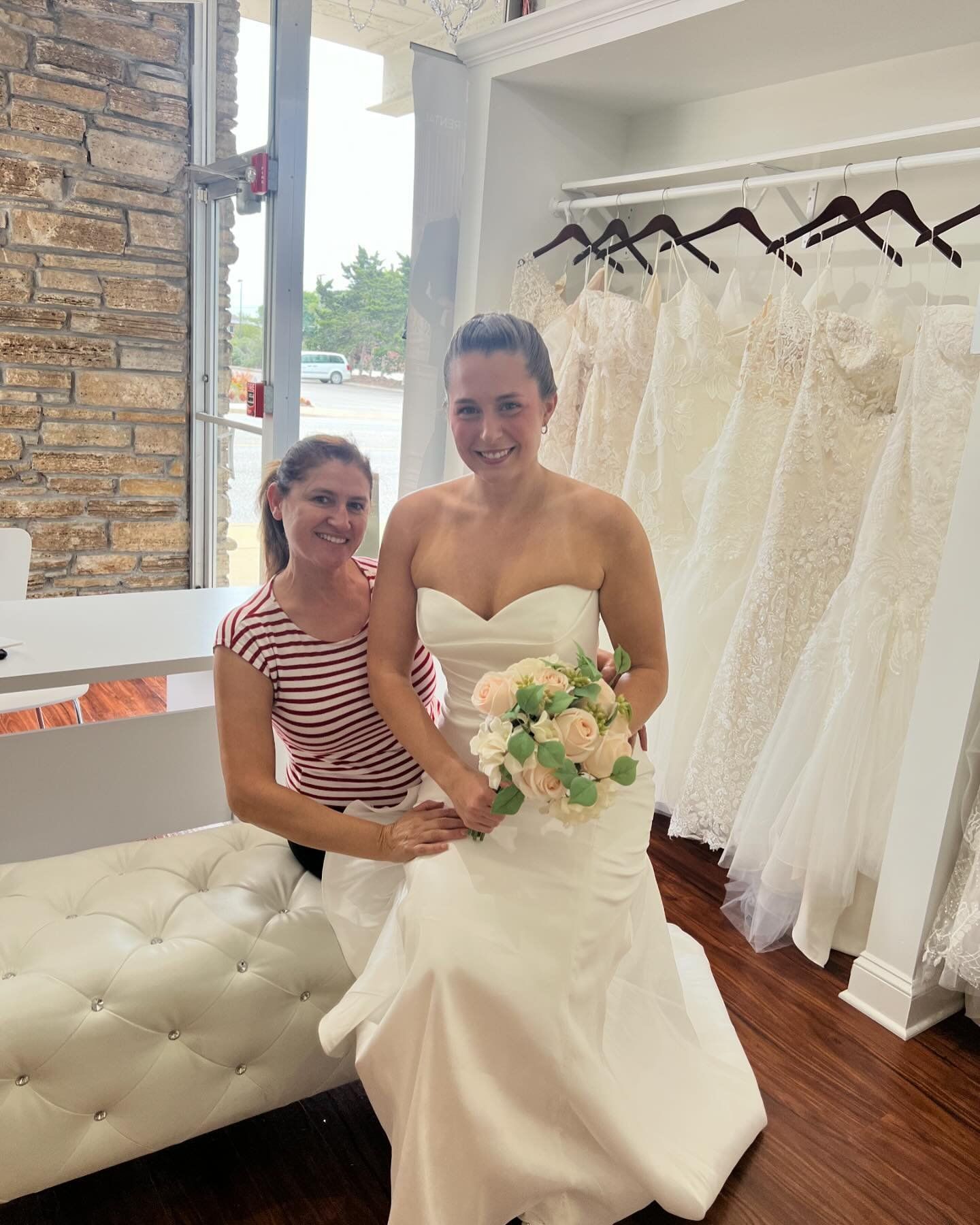 Woman trying on a wedding dress with an assistant in a bridal shop. The bride holds a bouquet.