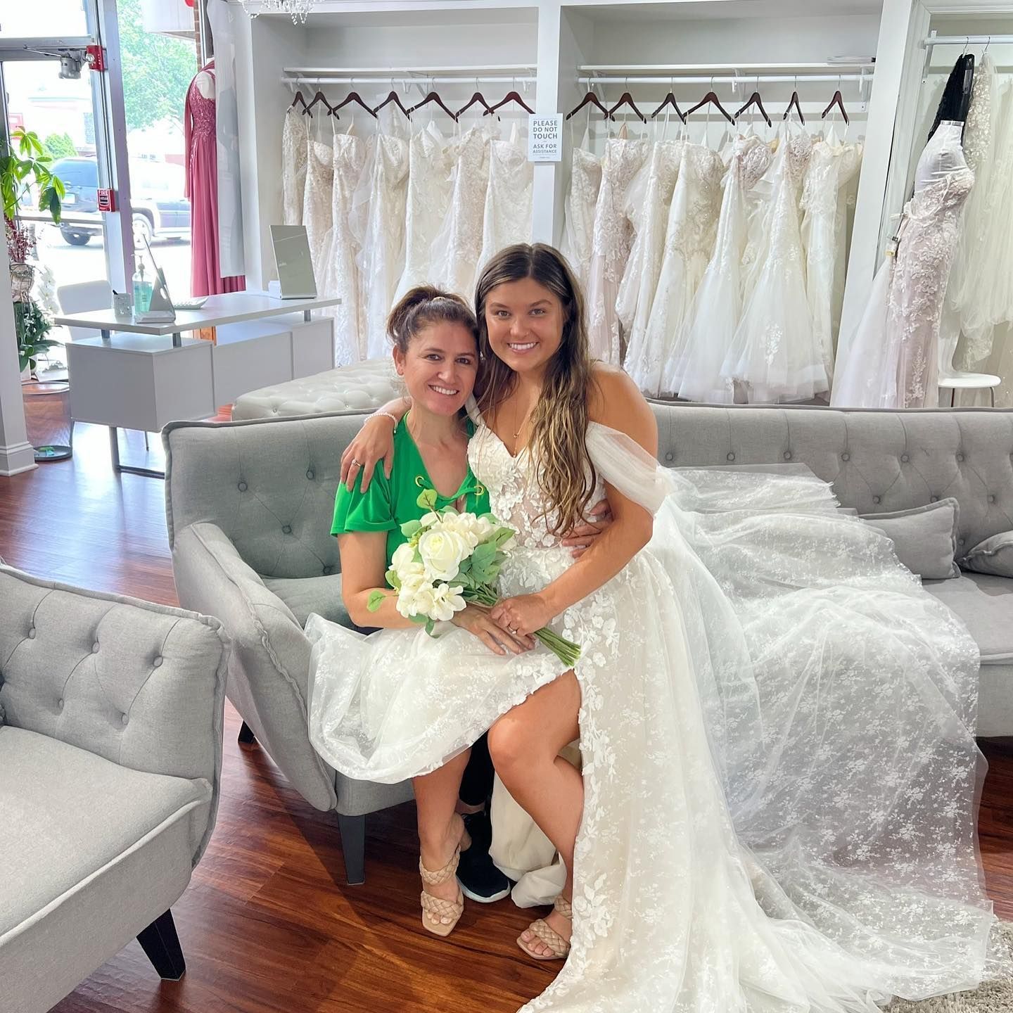 Two women smiling in a bridal shop, one in a wedding dress, the other holding flowers.