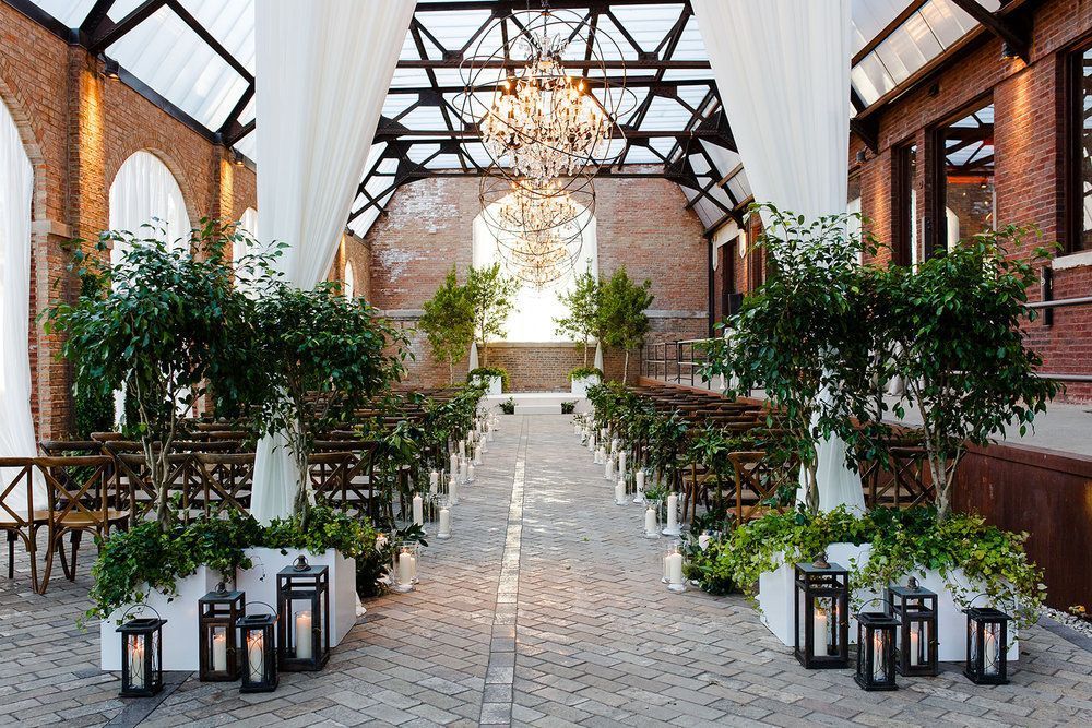 Indoor wedding ceremony setup, aisle lined with plants and lanterns, with a chandelier hanging overhead.