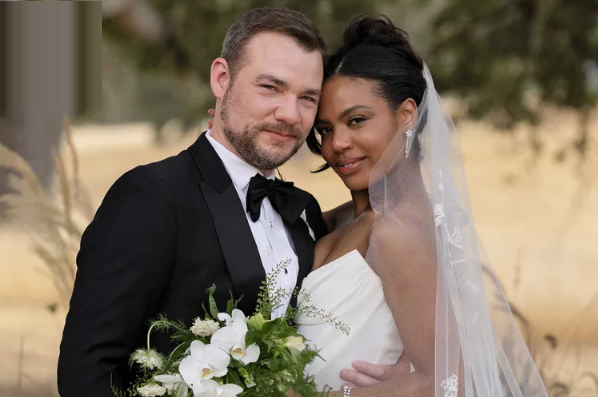 Couple in wedding attire embrace, holding flowers. Man in tuxedo, woman in strapless gown, outdoors.