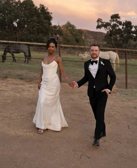 A bride and groom hold hands walking outdoors near horses and a fence at sunset.