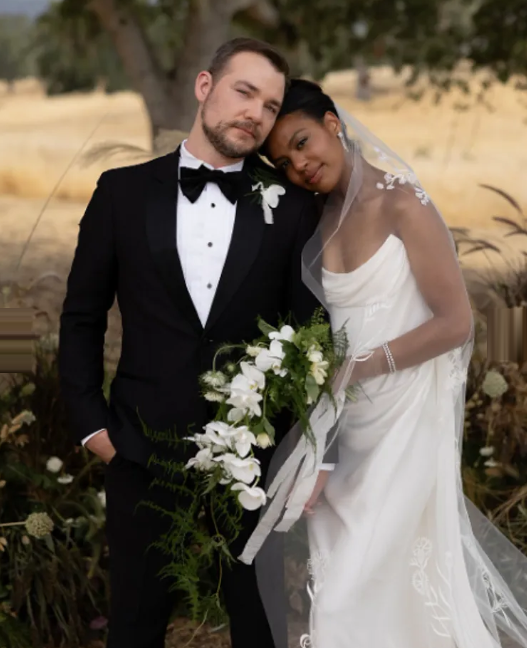 Newlyweds pose together; groom in a tuxedo, bride in a white gown with a veil and holding a bouquet.
