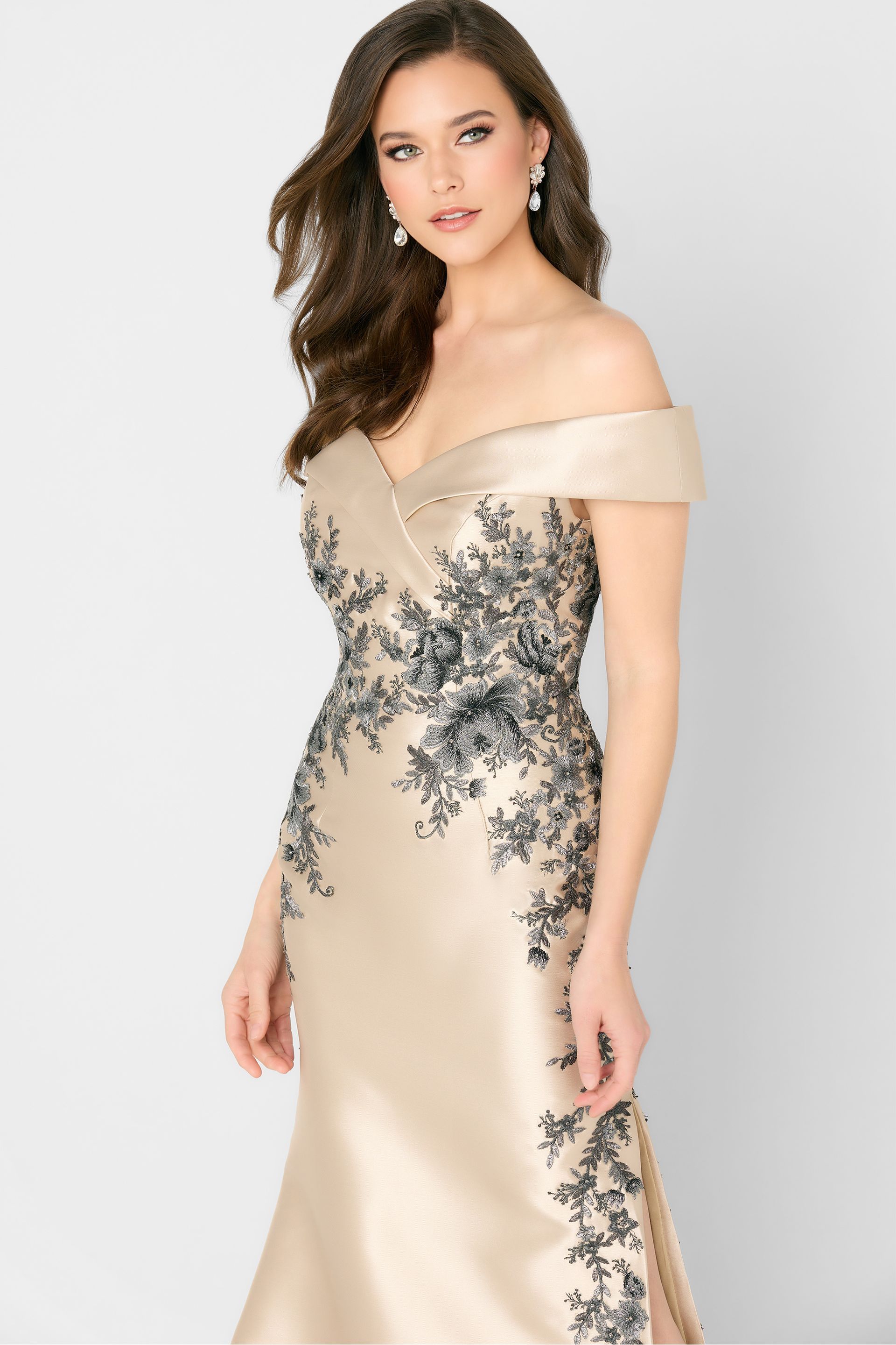 Woman in a beige off-shoulder gown with black floral embroidery, posing against a light background.