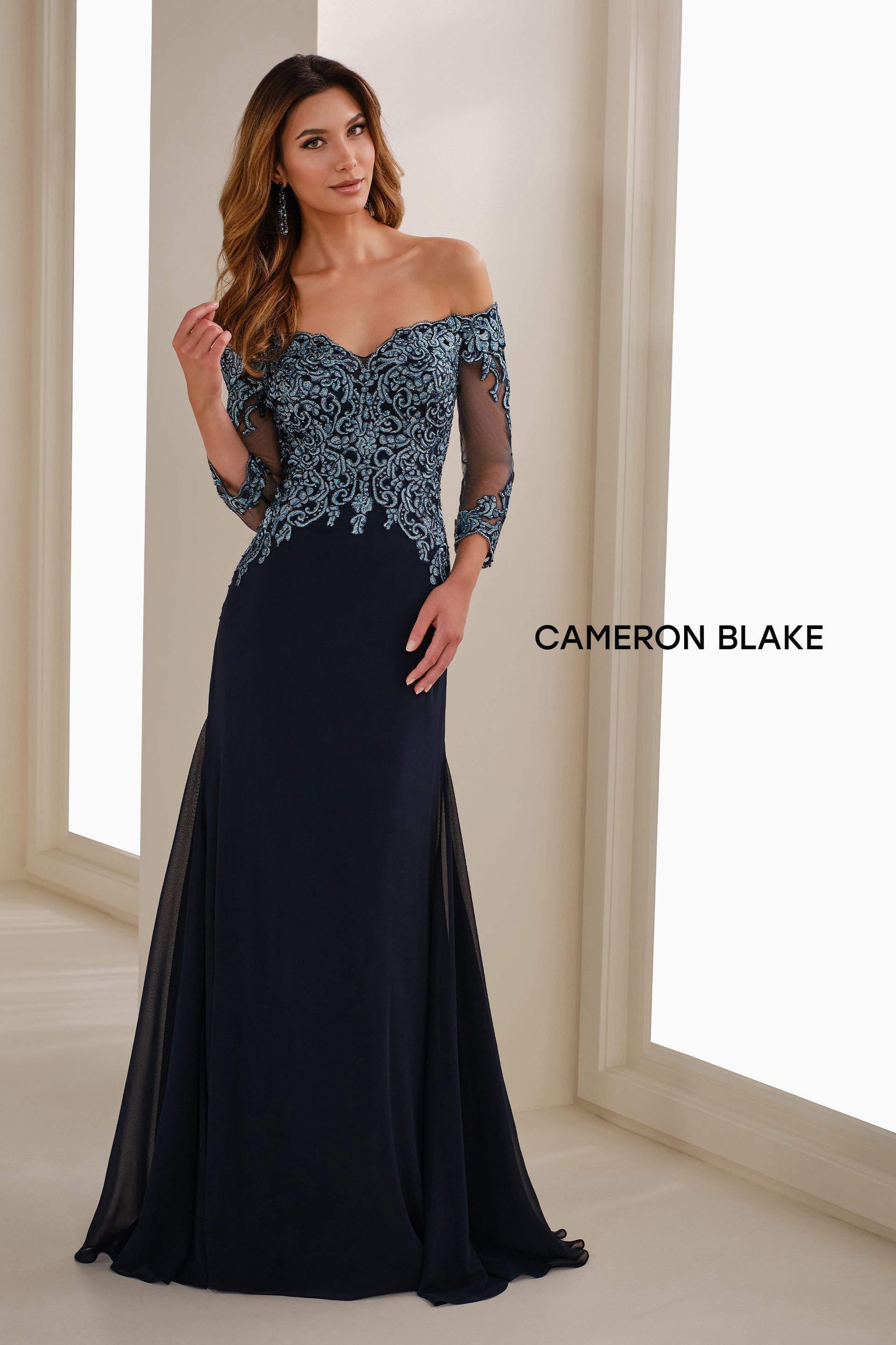 Woman in formal navy blue off-the-shoulder gown with sequined bodice, standing in a well-lit room.