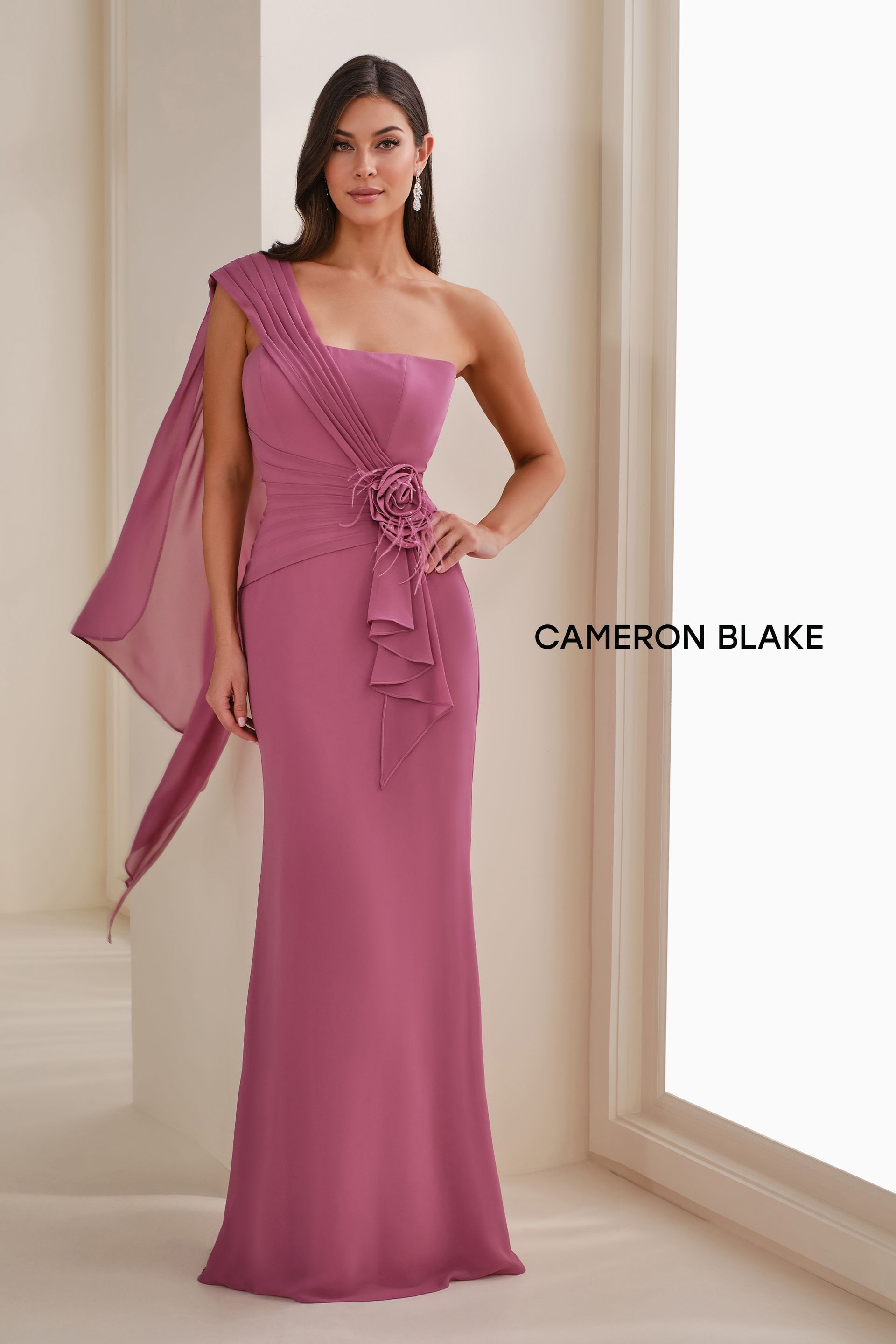 Woman wearing a mauve one-shoulder evening gown with cape detail and floral embellishment.