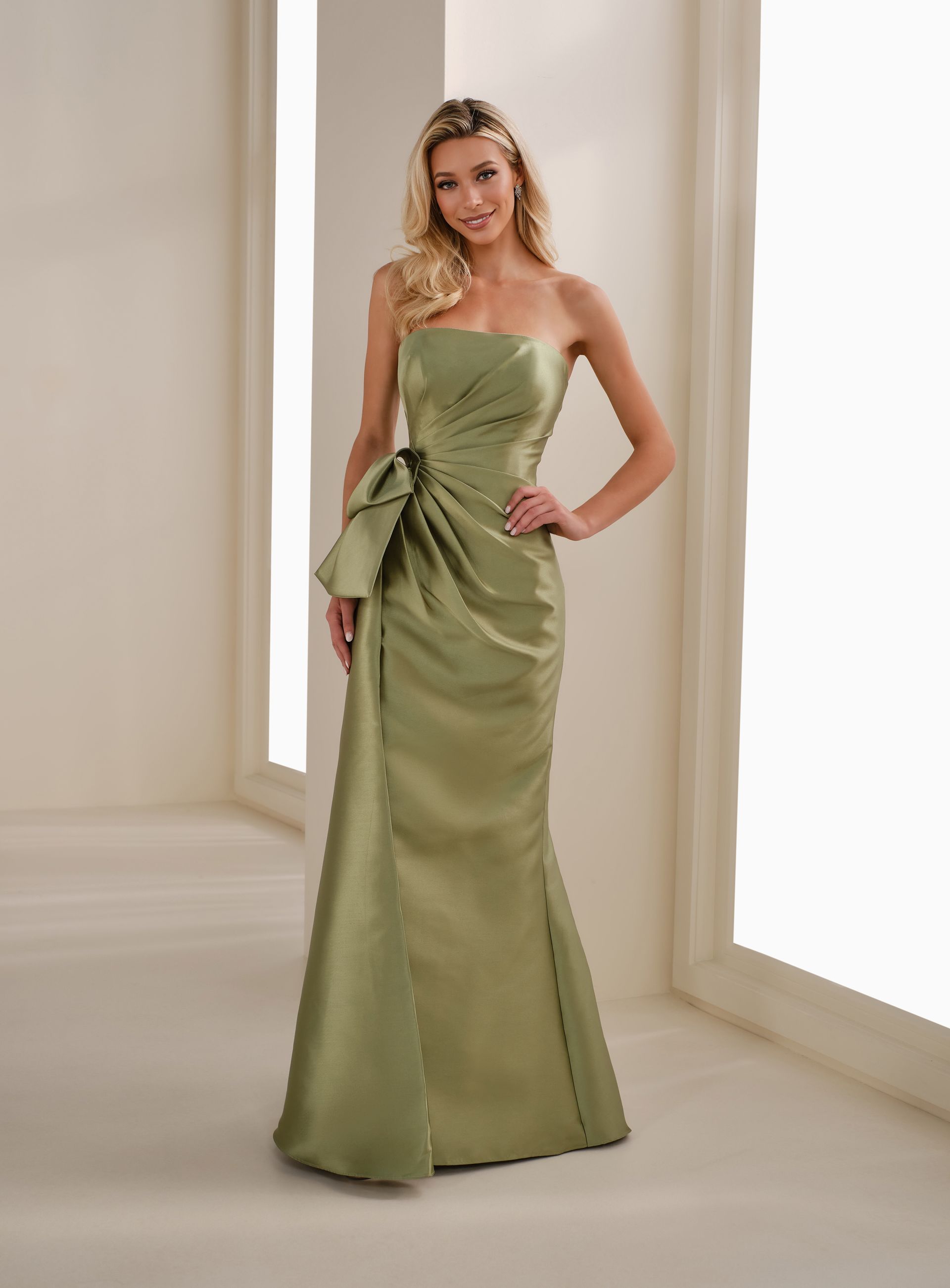 Woman in an olive green strapless gown standing in a bright studio with cream curtains.