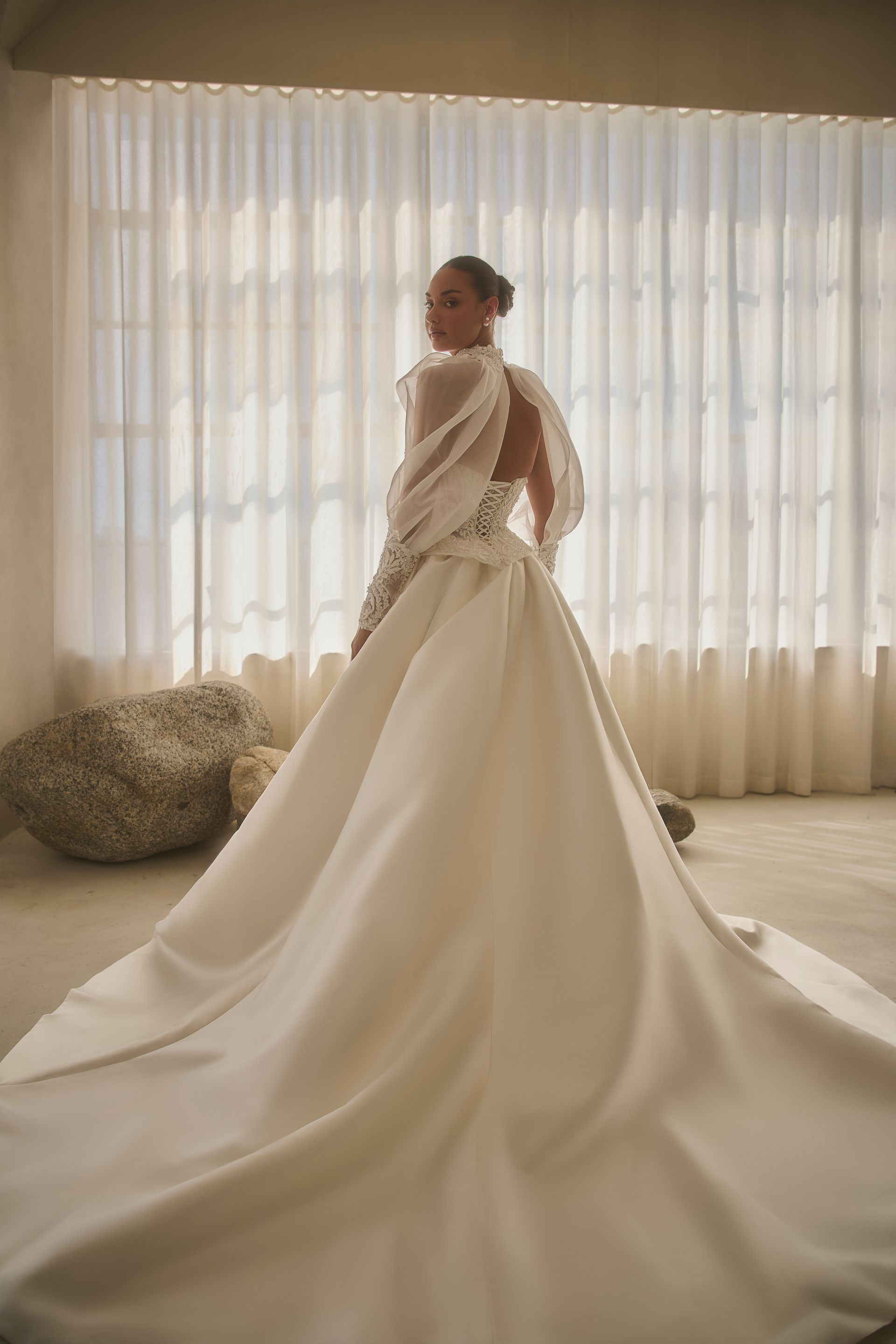 Woman in white wedding dress with open back, standing by window with long train.