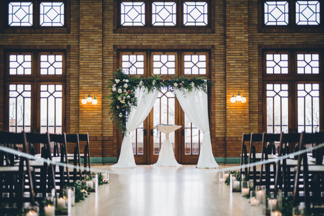 Wedding ceremony setup in a grand hall with floral arch, draped fabric, and candlelit aisle.