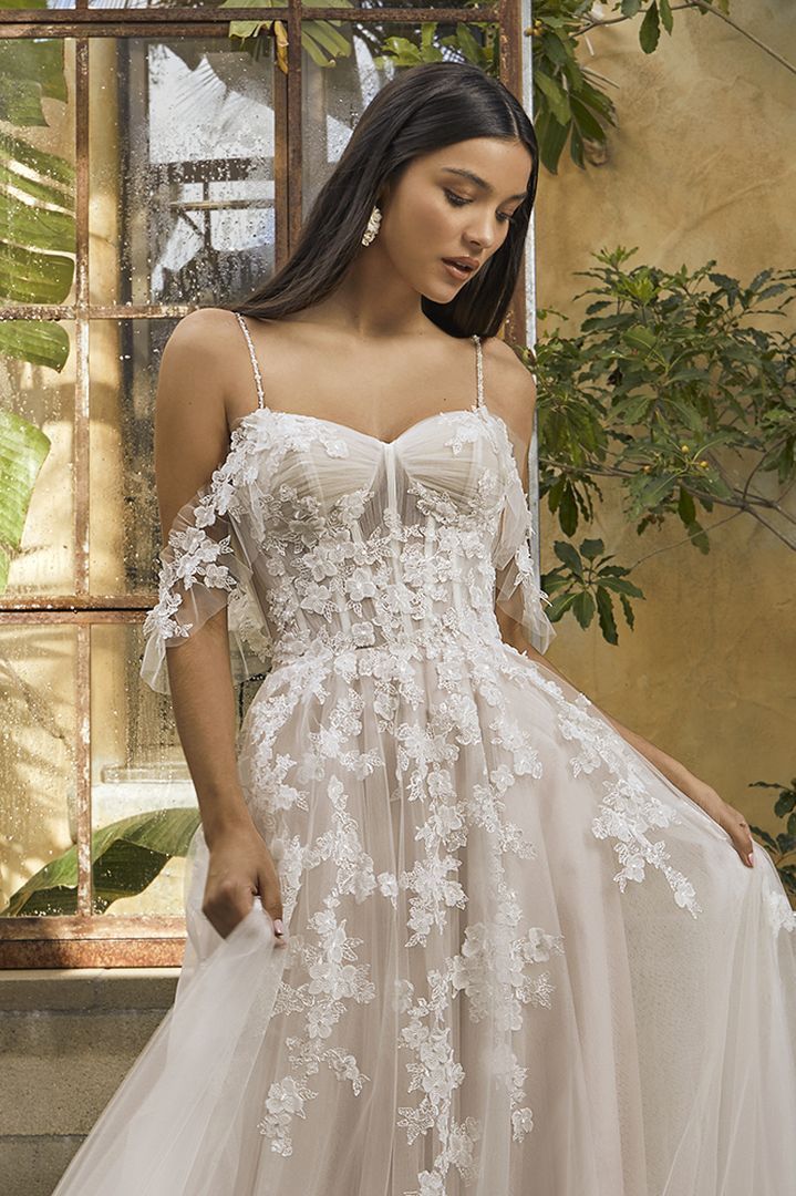 Woman in an off-the-shoulder, floral lace wedding gown. Outdoors, by a rustic window.