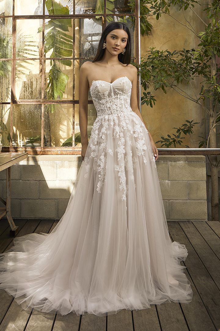 Strapless wedding dress with floral embellishments; woman stands on wooden deck.