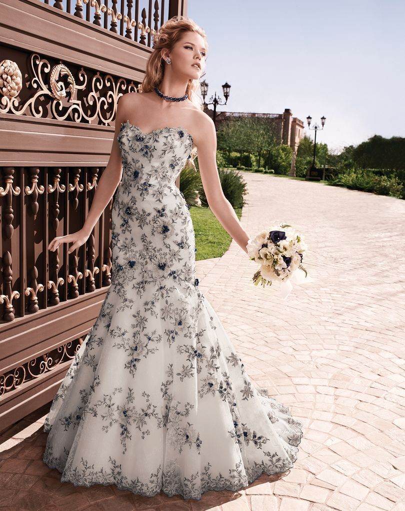 Woman in a white strapless wedding dress with blue floral embroidery, holding flowers, standing near a gate.