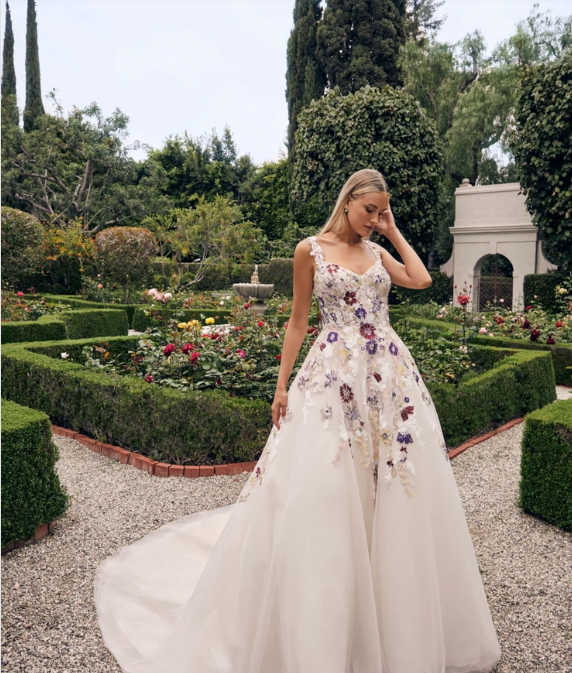 Woman in floral wedding gown poses in a garden, with flowers and hedges.