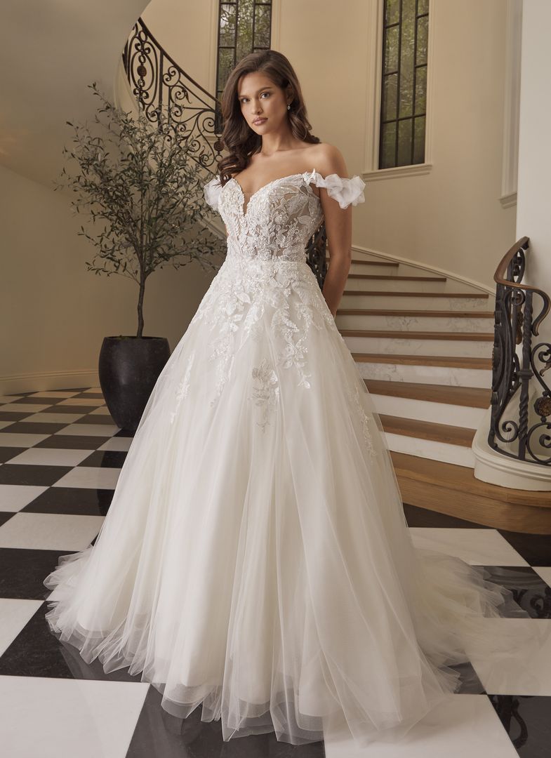 Woman in an off-the-shoulder wedding gown standing on checkered floor near a staircase.