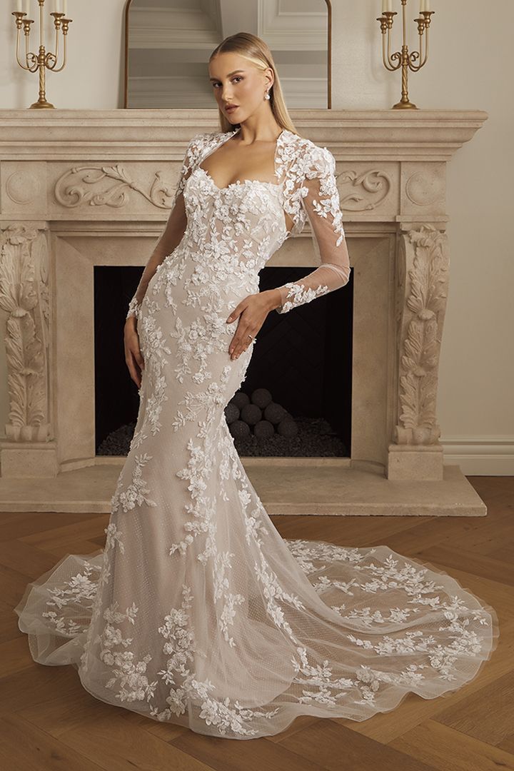 Woman in white lace wedding dress with long train, leaning on a fireplace.