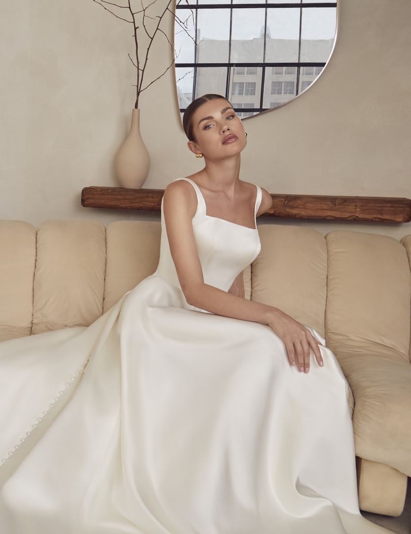 Woman in white satin wedding dress sitting on a beige couch, posing.