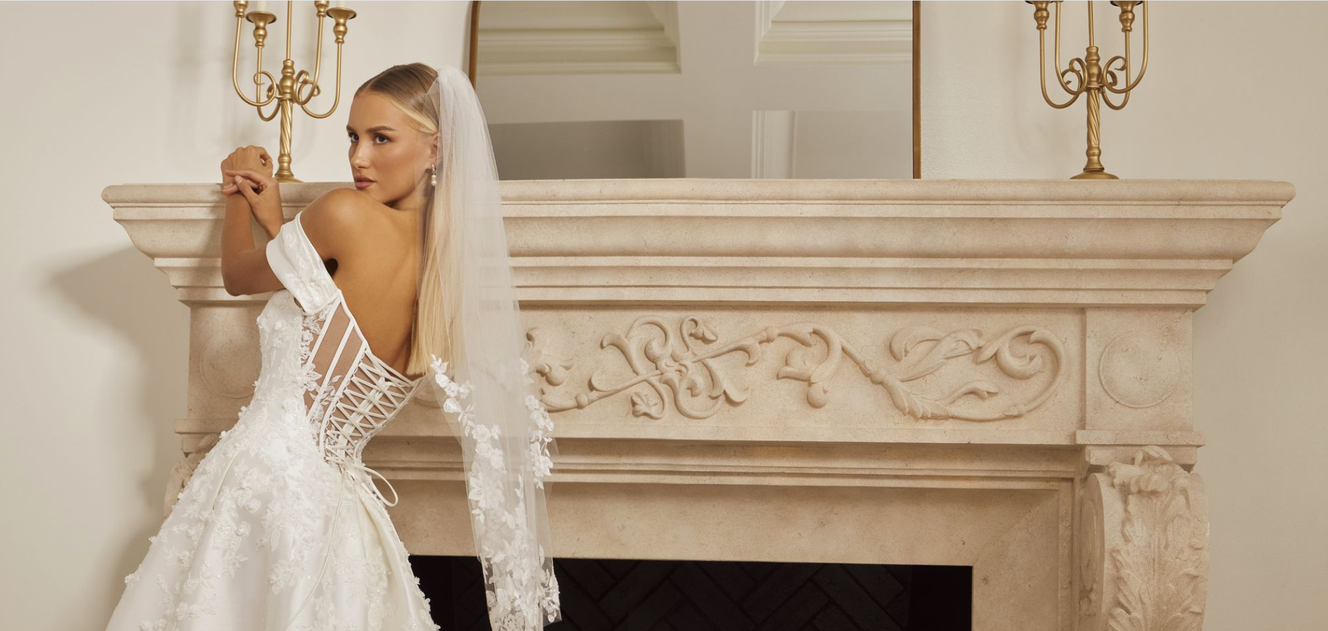 Bride in a white gown and veil leans near a fireplace with golden candelabras.