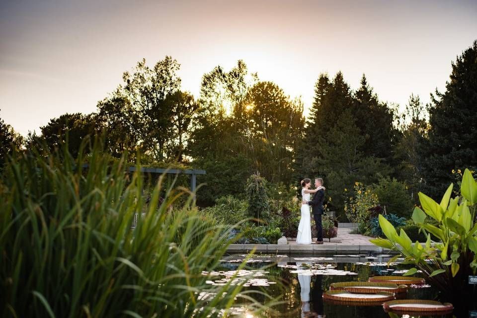Couple standing on a stone path by a pond at sunset, surrounded by greenery.