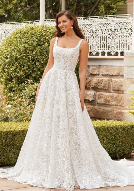 Woman in a white lace wedding gown, smiling, standing outside in front of greenery.