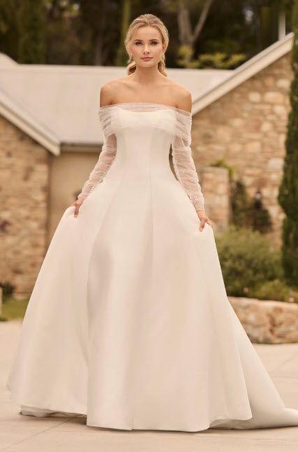 Woman in a white off-the-shoulder wedding dress with long, sheer sleeves, standing outside.