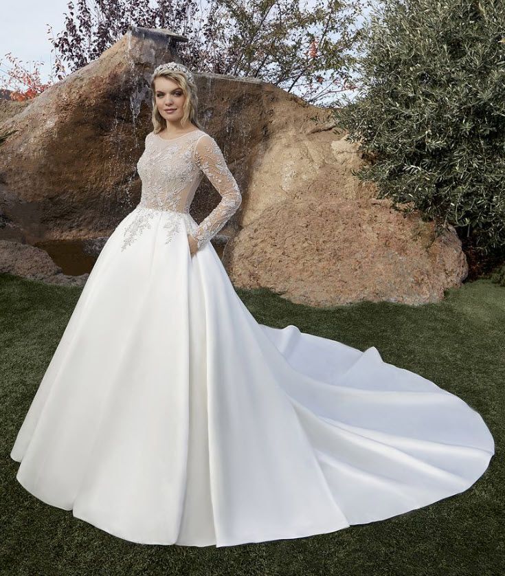 Bride in a white satin ball gown with a beaded bodice, long sleeves, and train, standing outside.