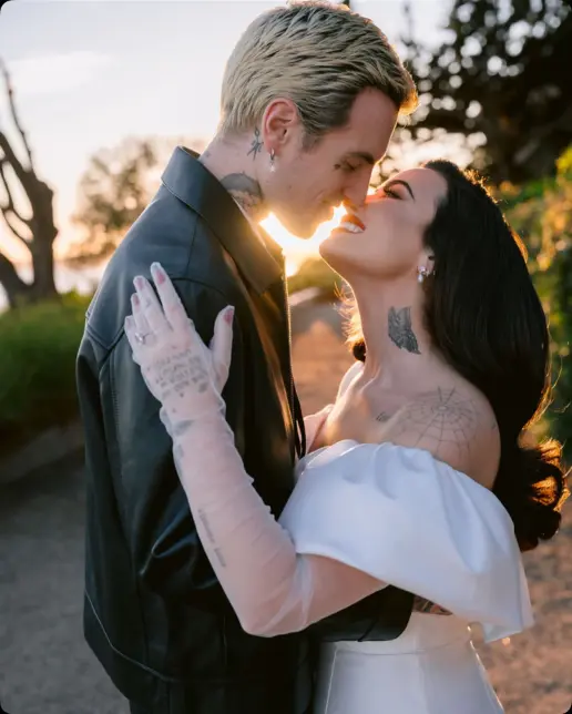 Couple embracing, about to kiss. Man in black jacket, blonde hair. Woman in white dress, dark hair. Golden sunlight.