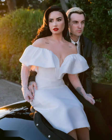 Demi Lovato in a white off-the-shoulder dress with a man, outdoors, posing on a car.